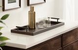 Rectangular dark brown tray with metal handles holding a gold cocktail shaker and two clear glass tumblers on a white countertop beside framed wall art.