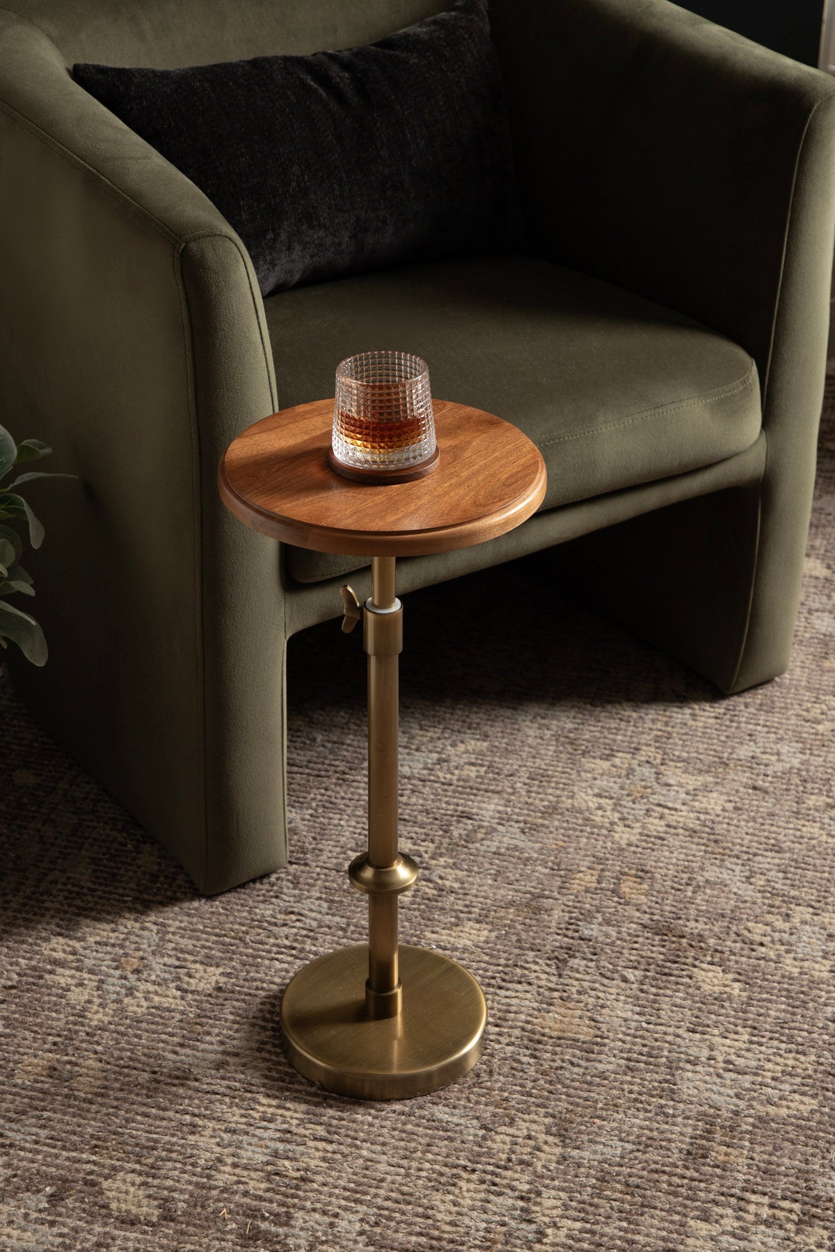 Round wood tabletop with a single glass of amber liquid on an adjustable brass stand beside an olive green armchair with a black cushion.