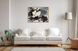 Abstract black, gray, and beige framed canvas hung above a white three-seat sofa with cushions, flanked by a potted plant on the left and a floor lamp on the right in a minimalist living room.