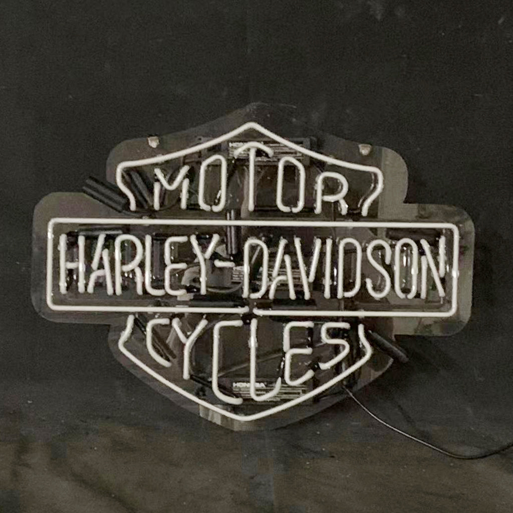White neon sign shaped like the Harley Davidson shield with the words MOTOR, HARLEY DAVIDSON, and CYCLES illuminated against a black background.