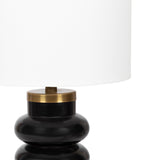 Table lamp with a bulbous black ceramic base topped by a brass-colored ring and a wide white fabric drum shade, shown against a white background.