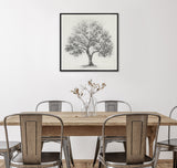 Framed charcoal-style drawing of a leafless tree with a single branch curving downward, hanging centered above a rustic wood dining table with metal chairs and glass vase.