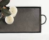 Rectangular dark hammered metal tray with a raised rim and a side handle, holding two white candles and green leaves, shown from above on a white background.
