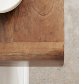 Close-up of a wooden table corner with a rounded edge, showing rich brown wood grain and slightly curved lines, set against a textured light floor.