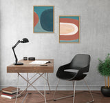 Two abstract framed art pieces with teal, rust, dark blue, and cream colors hang above a modern wooden desk and black chair on a textured gray wall with a potted plant nearby.