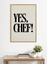 Minimalist wall art with large black serif text reading 'YES, CHEF!' on a light gray background in a simple wooden frame above a wooden table with books and two potted plants.