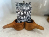 Two wooden teak kuksa mugs with hanging holes, arranged side by side on a white surface holding a black and white bag of Rise Up Organic Coffee House Roast.
