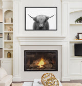 Black and white framed canvas of a Highland cow with long horns and shaggy hair, hung above a white fireplace with a burning fire inside.