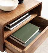 Light brown wooden side table with rounded edges, featuring a shelf holding closed black and white notebooks and an open drawer containing two stacked green notebooks.