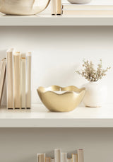 Gold-colored metal candle bowl with a wavy rim and white candle inside, placed on a white shelf between cream-colored books and a small white vase with dried flowers.
