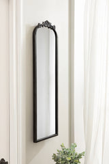 Tall, narrow arched wall mirror with a black frame and ornate floral decoration at the top, mounted on a light-colored wall near sheer white curtains and a small green plant below.