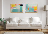 Two abstract framed canvas artworks with pastel blocks of teal, pink, yellow, and gray, hung side by side above a white cushioned sofa in a minimalist living room.