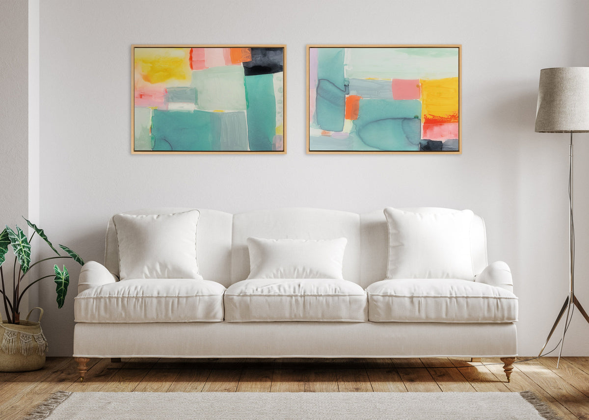 Two abstract framed canvas artworks with pastel blocks of teal, pink, yellow, and gray, hung side by side above a white cushioned sofa in a minimalist living room.