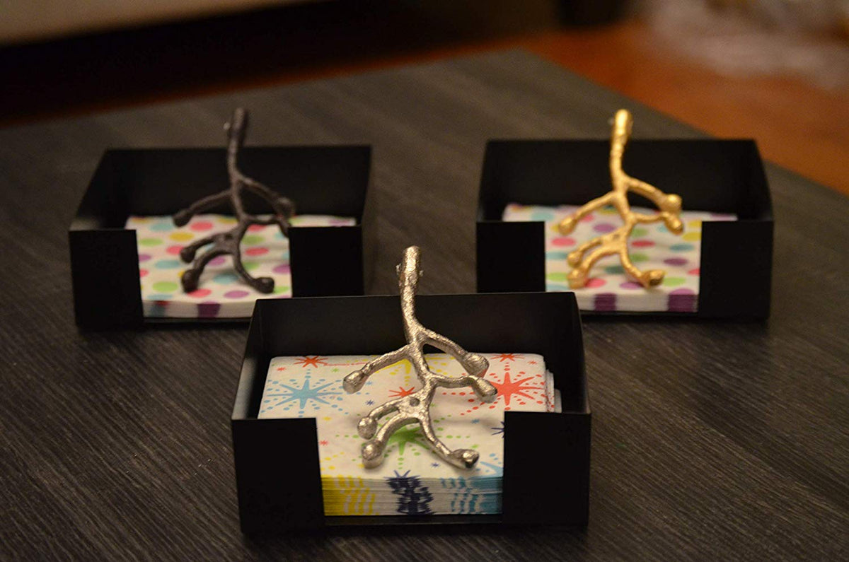 Three black square napkin holders each containing colorful napkins and topped with metallic branch-shaped accents in silver, gold, and dark bronze finishes.