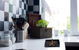 Black napkin holder on white kitchen countertop with a golden olive bud branch design, near a window and checkered black-gray tiled wall.