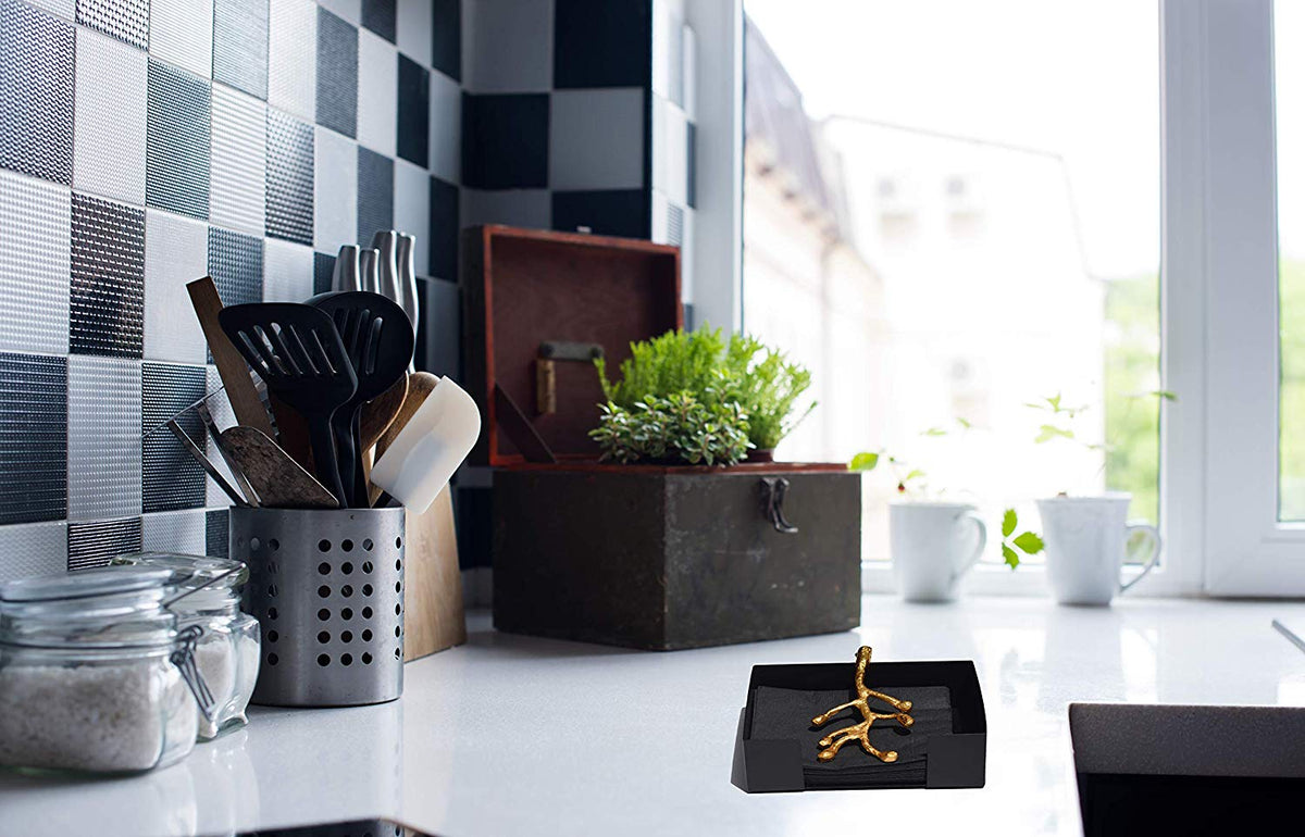 Black napkin holder on white kitchen countertop with a golden olive bud branch design, near a window and checkered black-gray tiled wall.