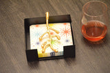 Black square napkin holder with a golden olive bud-shaped handle holding colorful star-patterned napkins, placed on a wooden table next to a glass of dark red liquid.