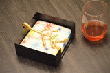 Black square napkin holder with a golden olive bud-shaped handle on patterned napkins, placed on a dark wood surface beside a glass of amber liquid.