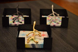 Three black square napkin holders each containing patterned napkins topped with metallic sculptures resembling branches in gold, silver, and dark bronze colors on a dark wood surface.