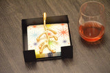 Black square napkin holder with a golden olive bud-shaped embellishment holding colorful star-patterned napkins, beside a glass of reddish drink on a woodgrain surface.