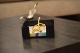 Black rectangular napkin holder with a golden olive branch-shaped handle holding patterned napkins, placed on a dark wood table with a silver leaf decor in the background.
