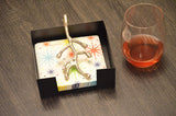 Square black napkin holder with silver branch-shaped handle holding colorful star-patterned napkins, next to a glass of reddish liquid on a dark wood surface.