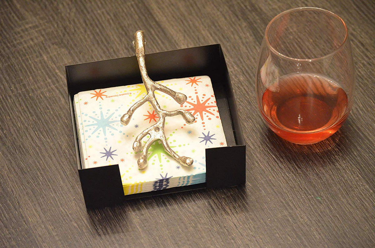 Square black napkin holder with silver branch-shaped handle holding colorful star-patterned napkins, next to a glass of reddish liquid on a dark wood surface.