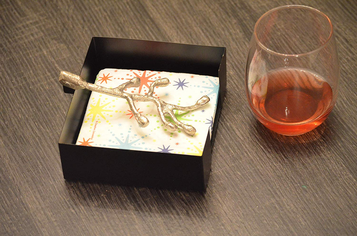 Silver branch-shaped napkin holder holding colorful star-patterned napkins inside a black square tray, next to a glass with amber liquid on a dark wood surface.