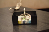 Black rectangular napkin holder containing patterned napkins with a silver branch-shaped napkin clip on top, placed on a textured gray surface.