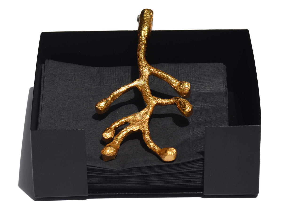 Matte black napkin holder containing black napkins with a gold branch-shaped decorative piece resting on top.