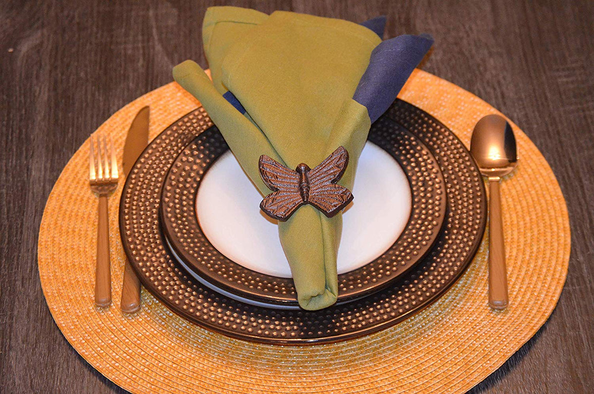 Green and blue folded napkin secured by a brown butterfly-shaped napkin ring on stacked textured dark plates with gold cutlery on a woven placemat.