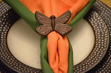 Antique bronze butterfly-shaped napkin ring holding folded orange and green cloth napkins on a white plate with a textured metallic rim.