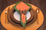 Dark wood butterfly-shaped napkin ring holding a folded green and orange napkin, placed on textured brown plates and woven round placemat with gold flatware.