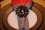 Black floral napkin ring with petal-shaped elements and a central pearl, holding a gray napkin on a white plate with a textured dark bronze charger