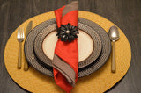 Table setting with layered brown textured plates on a round yellow woven placemat, featuring a red and brown napkin held by a black floral napkin ring with a pearl center.