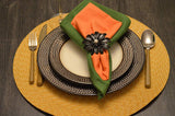 Table setting with textured dark brown plates on a woven yellow placemat, featuring a folded green and orange napkin secured by a black floral napkin ring.