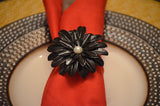 Black floral napkin ring with layered petals and a pearl center holding a folded red napkin on a white plate with a textured dark rim on a wooden surface.