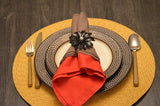 Black floral napkin ring with a pearl center holds folded red and brown napkins on a beige plate with a brown dotted border, placed on a round woven yellow placemat.