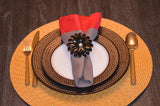 Table setting with a black floral napkin ring holding a red and gray folded napkin, placed on brown textured plates and a woven round placemat.