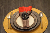 Black flower-shaped napkin ring with a central pearl securing brown and red folded napkins on a textured brown metal plate over a yellow woven placemat