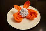 White floral napkin ring with a central pearl detail securing an orange fabric napkin folded into two pointed shapes on a white round plate against a dark background.