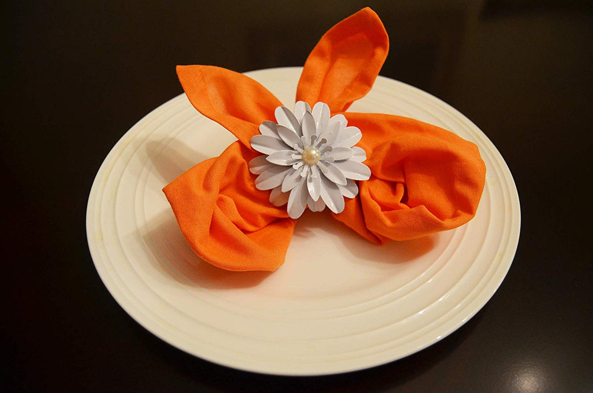 White floral napkin ring with a central pearl detail securing an orange fabric napkin folded into two pointed shapes on a white round plate against a dark background.