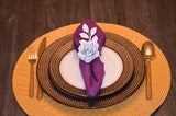 Dark brown ceramic plates with textured edges layered on a circular woven placemat, holding a purple cloth napkin folded with a white rose-shaped napkin ring on top.