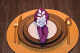White paper rose napkin ring holding a purple cloth napkin on a patterned brown dinner plate with woven placemat beneath on a wooden table with gold cutlery.
