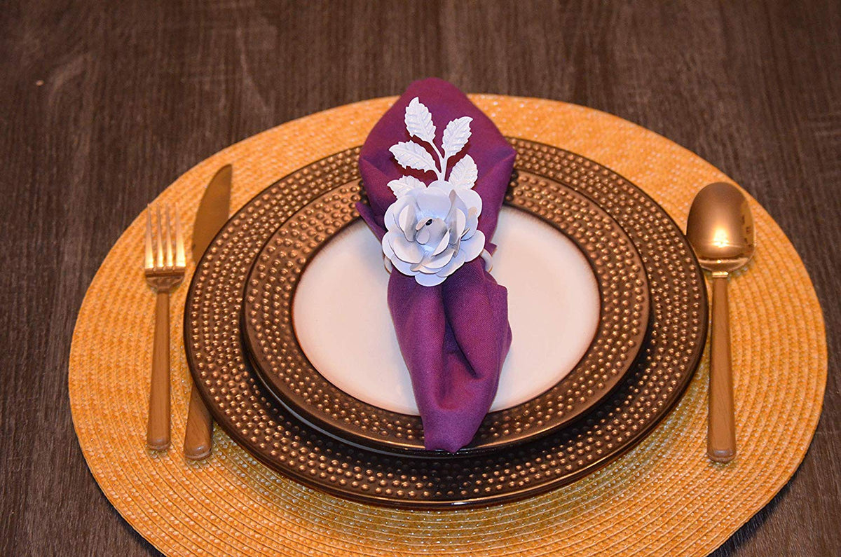 White paper rose napkin ring holding a purple cloth napkin on a patterned brown dinner plate with woven placemat beneath on a wooden table with gold cutlery.