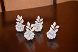 Set of four white rose napkin rings with detailed leaves, displayed on a dark reflective surface creating soft reflections beneath each piece.