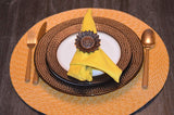 Yellow fabric napkin held by a brown sunflower-shaped napkin ring, placed on nested white and brown dotted ceramic plates with gold cutlery on a round woven placemat.