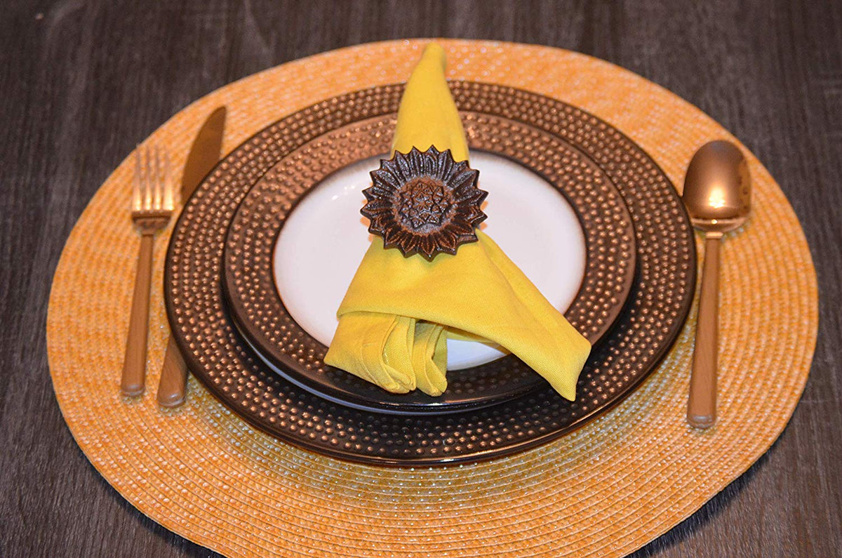 Yellow fabric napkin held by a brown sunflower-shaped napkin ring, placed on nested white and brown dotted ceramic plates with gold cutlery on a round woven placemat.