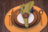 Green and blue folded napkin secured with a sunflower-shaped antique napkin ring on a white plate with a dark brown dotted plate underneath, placed on a round woven placemat with gold flatware on a wooden table.
