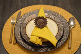 Table setting featuring a yellow napkin folded through a dark brown sunflower-shaped napkin ring on textured silver and black plates with gold flatware and a woven placemat